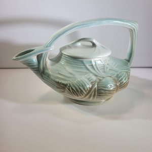 May include: A light blue ceramic teapot with a curved handle and lid. The teapot has a unique design with leaf-like patterns and a spout. The teapot is approximately 25 cm long and 15 cm tall.