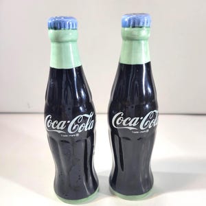 K&ouml;nnte beinhalten: Zwei Salz- und Pfefferstreuer in Form von Coca-Cola-Flaschen aus Keramik. Die Flaschen sind schwarz mit wei&szlig;em "Coca-Cola"-Schriftzug und einem gr&uuml;nen Hals. Die Flaschendeckel sind blau. Im Vintage-Stil.