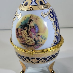 May include: A decorative egg-shaped trinket box with a hinged lid. The white porcelain egg features gold and blue floral patterns, a central painted scene, and rests on three gold-tipped feet. The box is trimmed with gold accents.