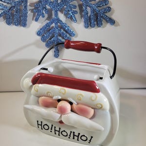 Puede incluir: Un recipiente de cerámica blanca con forma de Papá Noel con asa y borde rojos. El recipiente tiene un asa de alambre negro y una nariz y bigote rosas. Las palabras "Ho! Ho! Ho!" están impresas en la parte delantera. Un copo de nieve azul brillante está en el fondo.
