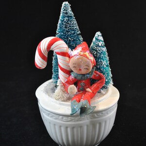 May include: A white ceramic Christmas music box featuring a red and white candy cane, a red elf figurine, and two green flocked Christmas trees.