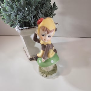 May include: A ceramic figurine of a seated elf wearing a yellow hat with a red dot, brown clothing, and tan shoes. The elf is perched on a green mushroom with white spots. A small, white planter with greenery is in the background.