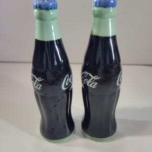 K&ouml;nnte beinhalten: Ein Paar Salz- und Pfefferstreuer aus Keramik in Form von Coca-Cola-Flaschen. Die Flaschen sind dunkelblau mit wei&szlig;em "Coca-Cola"-Schriftzug. Die Deckel sind hellgr&uuml;n mit blauen Kappen.