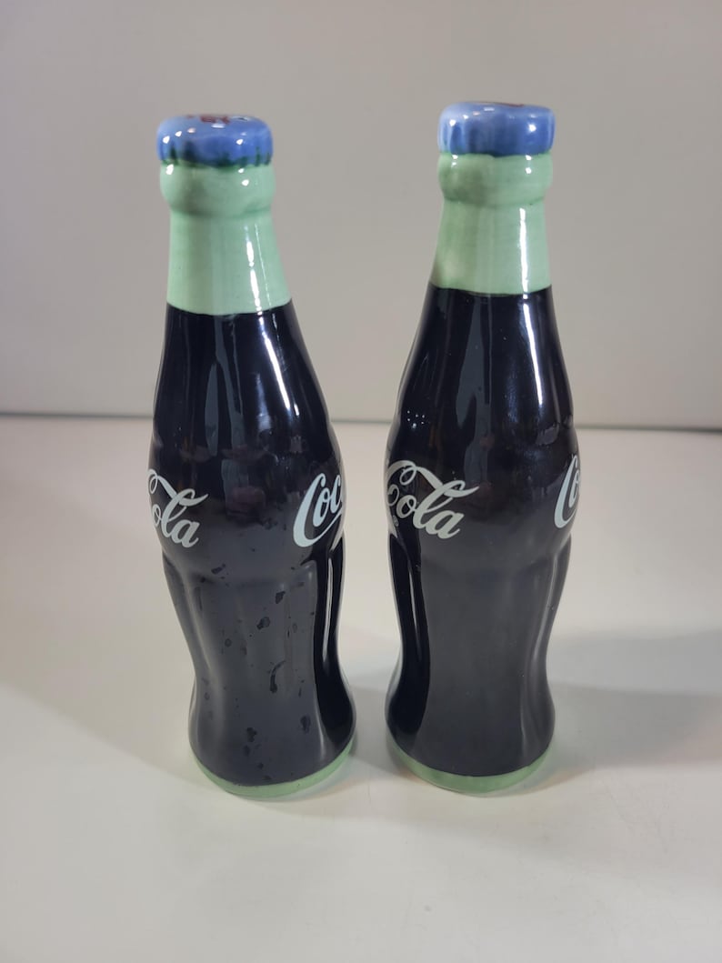 K&ouml;nnte beinhalten: Ein Paar Salz- und Pfefferstreuer aus Keramik in Form von Coca-Cola-Flaschen. Die Flaschen sind dunkelblau mit wei&szlig;em "Coca-Cola"-Schriftzug. Die Deckel sind hellgr&uuml;n mit blauen Kappen.