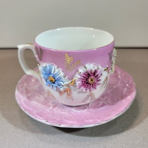 ピンク、黄色、青の立体的な花が描かれた美しいピンクのカップとソーサー、エンボス加工のソーサー、金色のアクセント