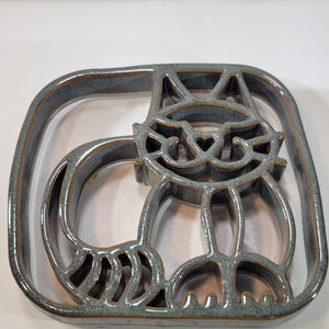 Puede incluir: Un salvamanteles de cerámica con forma de gato en un marco cuadrado. El salvamanteles es de color azul grisáceo apagado con un acabado brillante y un borde marrón. El diseño del gato presenta ojos en forma de corazón y una cola enroscada. Para la cocina.