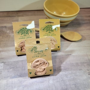 Puede incluir: Tres prensas para galletas de color marrón con un diseño floral. Cada prensa está empaquetada en una caja de cartón marrón con un logotipo verde que dice "Shaker Hearth simply good things cookie press".
