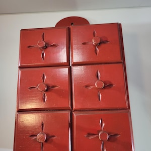 Könnte beinhalten: Ein rechteckiger Wand-Organizer aus Holz mit sechs quadratischen Schubladen in zwei Spalten. Jede Schublade ist dunkelrot lackiert und verfügt über einen runden Knopf in der Mitte, umgeben von vier geschnitzten blattförmigen Vertiefungen.