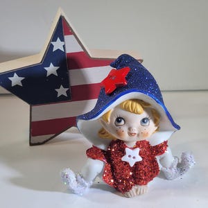 May include: A patriotic figurine of a seated elf with a red glittery top and a blue glittery hat with a red star. The elf has blonde hair and white gloves. A wooden star with a red, white, and blue design is in the background.