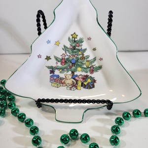 Puede incluir: Un plato de cerámica blanco en forma de árbol de Navidad con un borde verde. El plato presenta una colorida ilustración de un árbol de Navidad decorado, regalos y un oso de peluche. Un collar de cuentas verdes está en primer plano.