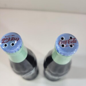 K&ouml;nnte beinhalten: Zwei Salz- und Pfefferstreuer aus Keramik in Form von Coca-Cola-Flaschen. Die Flaschen sind schwarz mit einem gr&uuml;nen Hals und einem blauen Deckel. Das Coca-Cola-Logo ist auf dem Deckel zu sehen.