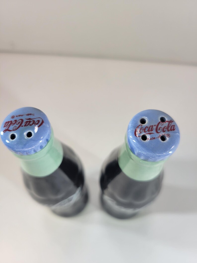 K&ouml;nnte beinhalten: Zwei Salz- und Pfefferstreuer aus Keramik in Form von Coca-Cola-Flaschen. Die Flaschen sind schwarz mit einem gr&uuml;nen Hals und einem blauen Deckel. Das Coca-Cola-Logo ist auf dem Deckel zu sehen.