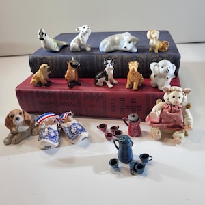 Puede incluir: Una colección de figuras de animales en miniatura de cerámica, incluyendo perros y una oveja, sobre dos libros apilados. El conjunto incluye un par de zuecos de madera pintados en blanco y azul, una cafetera roja pequeña con tazas y una cafetera azul.