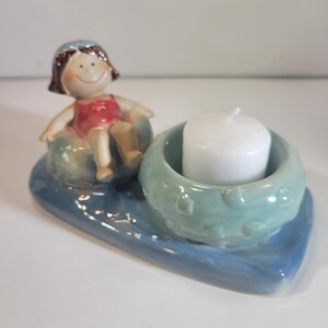 May include: A ceramic candle holder featuring a smiling figurine in a red swimsuit, seated on a blue and green float. A white candle sits in a teal holder, all on a blue base. The design is whimsical and decorative.