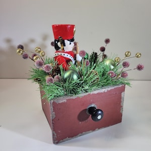 Puede incluir: Un cajón de madera vintage con un pomo negro, decorado con una escena navideña. Una figurita de soldado de juguete rojo y blanco está rodeada de ramas de pino verdes, bayas rojas y adornos verdes y dorados.