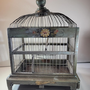 Puede incluir: Jaula para pájaros vintage con acabado pintado envejecido. La jaula presenta un diseño floral decorativo y una estructura de alambre metálico. La jaula tiene una base cuadrada y una parte superior abovedada, con un pináculo en el ápice.