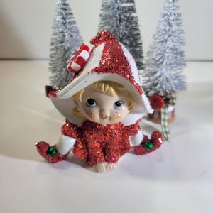May include: A ceramic elf figurine with blonde hair, wearing a red glittery outfit and a matching wide-brimmed hat topped with a red and white peppermint candy accent. The elf has green jingle bells on its shoes and is positioned in front of three small, flocked artificial pine trees.