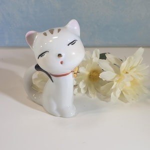 May include: A white ceramic cat figurine with closed eyes, gold accents, and a red collar. The cat has a black tail and is next to a bouquet of white flowers. The figurine is on a white surface with a light blue background.