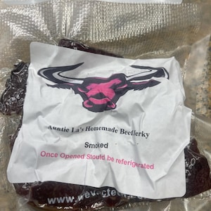 May include: A vacuum-sealed package of dark brown beef jerky with a white label featuring a black and pink bull head logo. The text reads Auntie La's Homemade Beef Jerky, Smoked, and Once Opened Should be refrigerated, with a website URL at the bottom.
