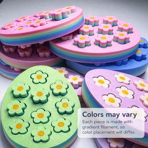 May include: A collection of colorful, flower-shaped molds in various pastel colors, including pink, blue, green, and purple. Each mold features multiple flower-shaped indentations with yellow centers. The molds have a rainbow-colored edge. Text on the image states "Colors may vary."