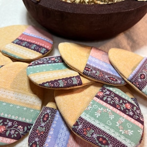 May include: A collection of yellow fabric coasters with floral and lace patterned accents in shades of pink, purple, and green. A dark brown wooden bowl filled with shredded paper sits in the background.