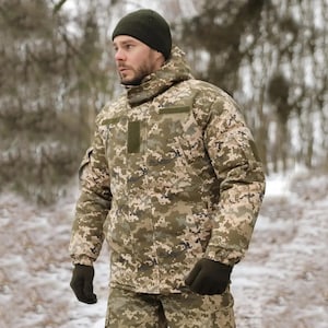 May include: A person wearing a camouflage jacket with a hood and a black beanie. The jacket has a zip and pockets. The person is standing in a snowy woodland.