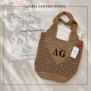 May include: A tan crochet mesh tote bag featuring a triangular patch with the black embroidered letters AG. The bag includes a white drawstring dust bag and a white rectangular tag with red string. A translucent plastic bag displays LAUREL CANYON WORKS.