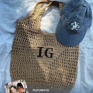 May include: A tan crochet tote bag with the letters IG printed in black on the front, accompanied by a faded blue baseball cap featuring a white NY logo. The image includes text stating Artisan Made, Hand painted in the USA, and Giggly Squad.