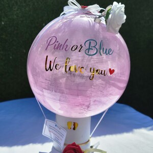Puede incluir: Globo de revelaci&oacute;n de g&eacute;nero rosa y azul con el texto "Pink or Blue We love you" y una rosa blanca en la parte superior. El globo est&aacute; sujeto a un soporte blanco con una rosa roja en la parte inferior.