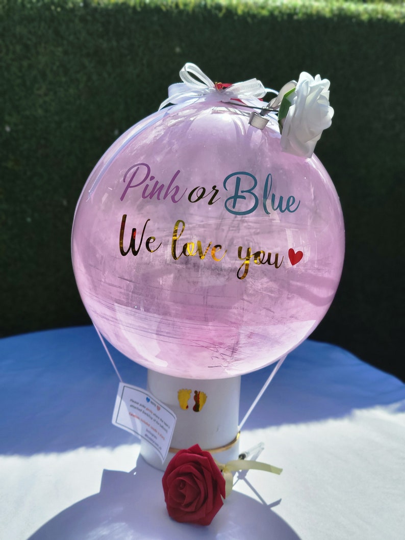 Puede incluir: Globo de revelaci&oacute;n de g&eacute;nero rosa y azul con el texto "Pink or Blue We love you" y una rosa blanca en la parte superior. El globo est&aacute; sujeto a un soporte blanco con una rosa roja en la parte inferior.