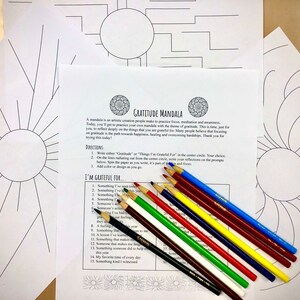 May include: A collection of mandala coloring pages with a gratitude theme, alongside a set of colored pencils. The pages feature intricate line drawings, including radiating sunburst designs and geometric patterns. The text on the page reads "Gratitude Mandala".