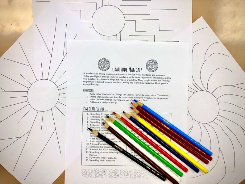 May include: A collection of mandala coloring pages with a gratitude theme, alongside a set of colored pencils. The pages feature intricate line drawings, including radiating sunburst designs and geometric patterns. The text on the page reads "Gratitude Mandala".