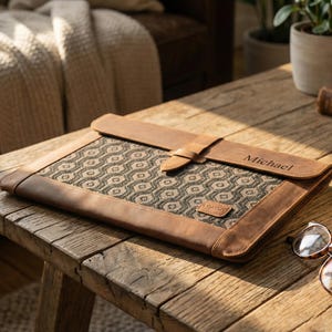 Könnte beinhalten: Eine braune Laptop-Hülle aus Leder mit einem geometrisch gemusterten Stoffmittelteil. Die Hülle hat einen Schnallenverschluss und der Name "Michael" ist auf die Klappe graviert. Ein "London"-Etikett ist sichtbar. Die Hülle liegt auf einem Holztisch.