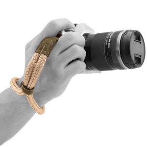 May include: A tan and brown camera wrist strap with a leather accent. The strap is wrapped around a person's wrist and is attached to a camera.