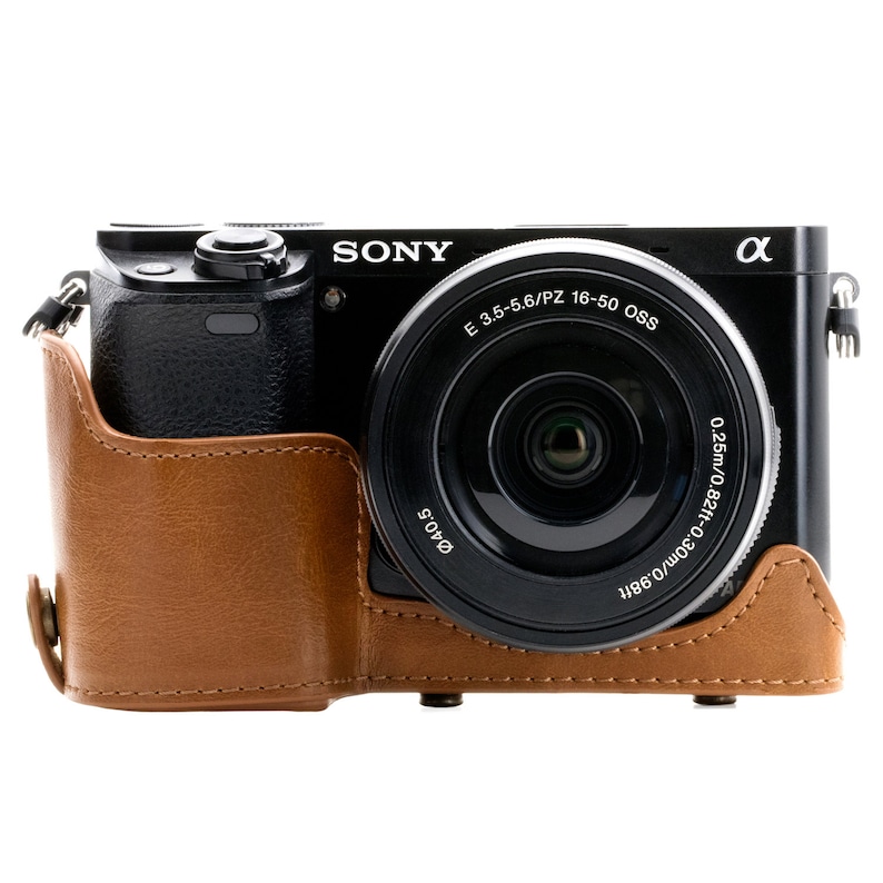 Pu&ograve; includere: Una fotocamera Sony Alpha nera con una custodia in pelle marrone e un obiettivo nero con il testo "E 3.5-5.6/PZ 16-50 OSS" e "0.25m/0.82ft-0.30m/0.98ft" stampato sull'obiettivo.