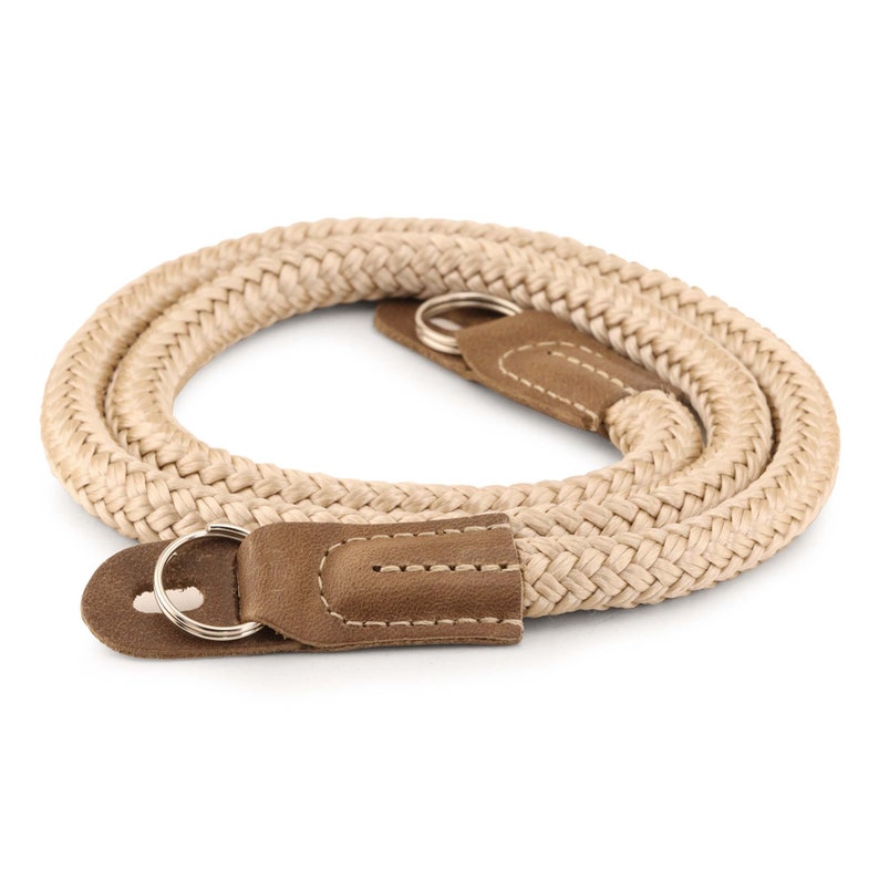 May include: A beige camera strap made of braided rope with brown leather accents and metal rings.