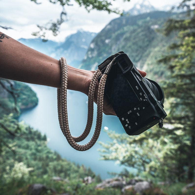 May include: A person holding a black camera with a tan camera strap. The camera has a variety of buttons and dials. The background is a blurry image of a lake and mountains.