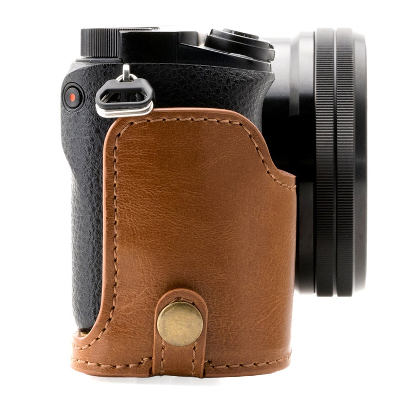 Pu&ograve; includere: Una custodia per fotocamera in pelle marrone con una fotocamera nera all'interno. La custodia ha un lembo che copre l'obiettivo e una cinghia che circonda la fotocamera.