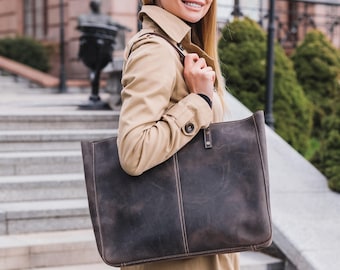 May include: A brown leather tote bag with a top handle and a zipper closure. The bag is worn by a person wearing a beige trench coat.