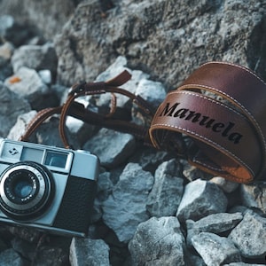 May include: A silver camera with a brown leather strap that says "Manuela" on it. The camera and strap are lying on a bed of grey rocks.
