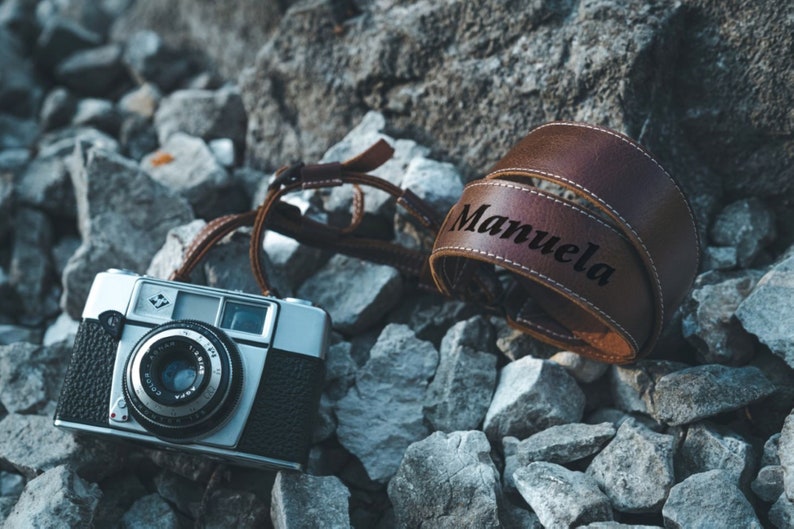 May include: A silver camera with a brown leather strap that says "Manuela" on it. The camera and strap are lying on a bed of grey rocks.