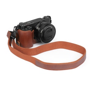 Può includere: Una custodia per fotocamera in pelle marrone con una tracolla abbinata. La custodia è progettata per adattarsi a una fotocamera mirrorless con un obiettivo.
