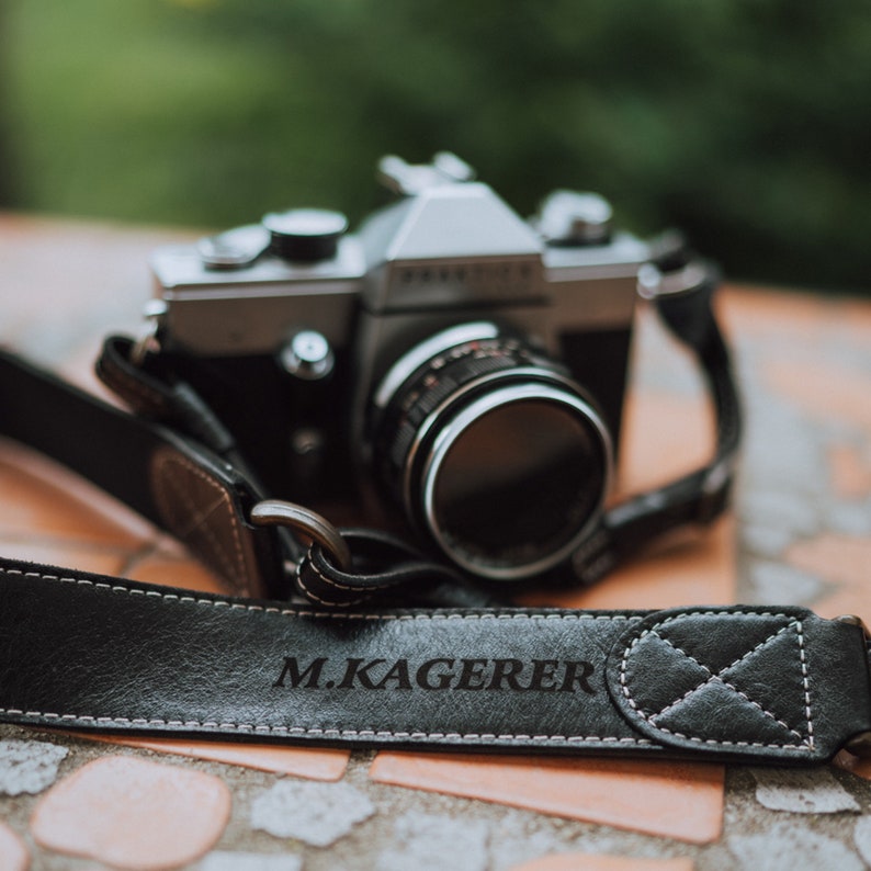 May include: A black leather camera strap with the text "M.KAGERER" embossed on it. The strap is attached to a silver and black film camera.