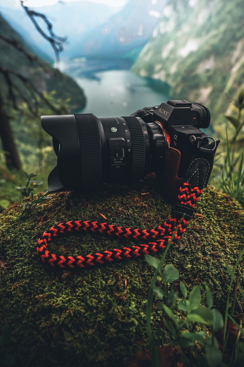 May include: A black camera with a large lens sits on a mossy rock. The camera has a red strap with a black and red braided design. The camera is pointed towards a body of water in the distance.