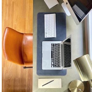 Op de afbeelding: Een bovenaanzicht van een werkplek met een bruine leren stoel, een laptop, een toetsenbord, een notitieblok en een gouden lamp. De laptop is open en het scherm toont een webpagina. Het toetsenbord ligt op een zwarte mat.