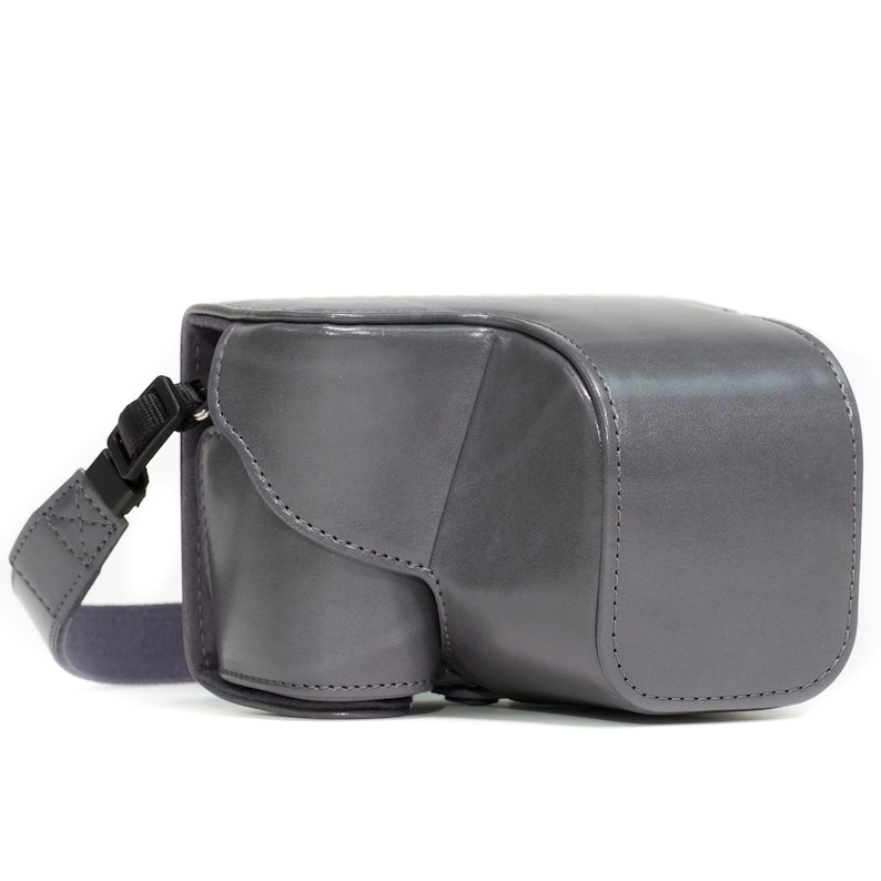 Pu&ograve; includere: Una borsa per fotocamera in pelle grigia con una tracolla. La borsa ha una patta che copre l'apertura e una chiusura con cerniera.
