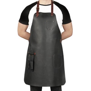 May include: A black leather apron with brown leather straps and multiple pockets. The apron is being worn by a person with a white t-shirt and black sleeves.