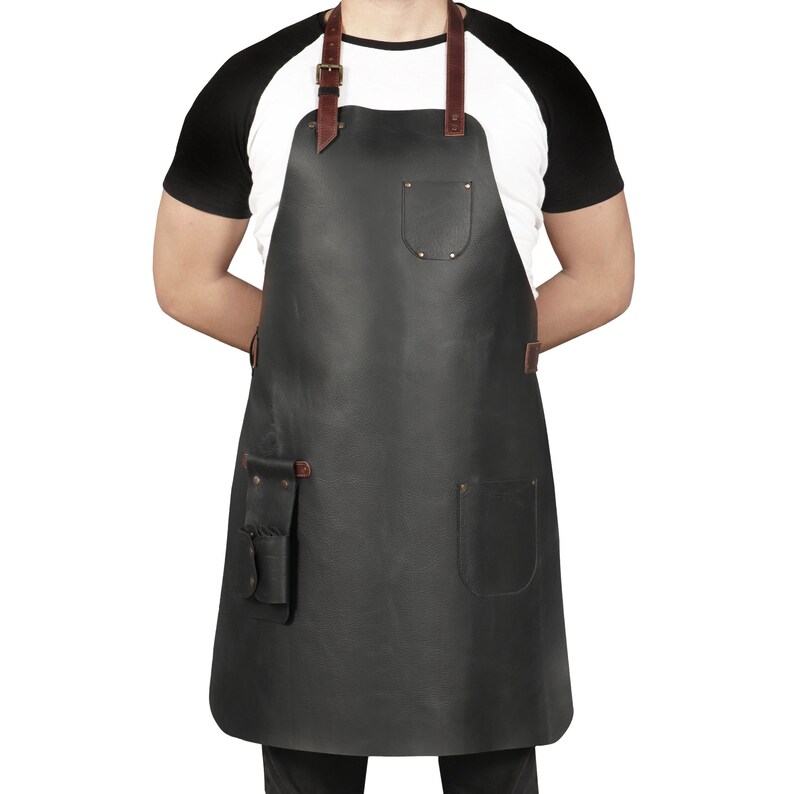 May include: A black leather apron with brown leather straps and multiple pockets. The apron is being worn by a person with a white t-shirt and black sleeves.