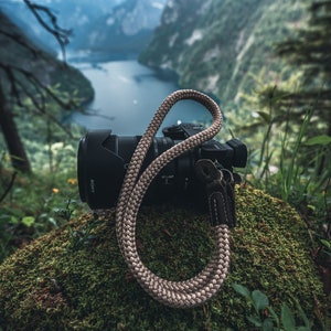 May include: A black camera with a lens and a beige camera strap. The camera is sitting on a bed of green moss.