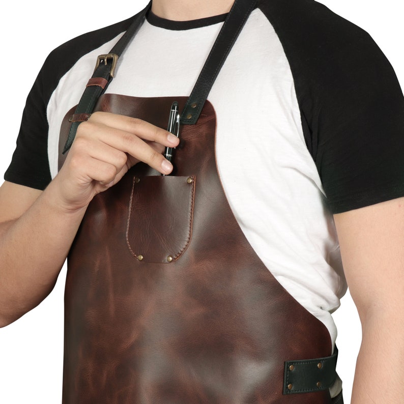May include: A brown leather apron with a black strap and a pocket on the front. A pen is in the pocket.